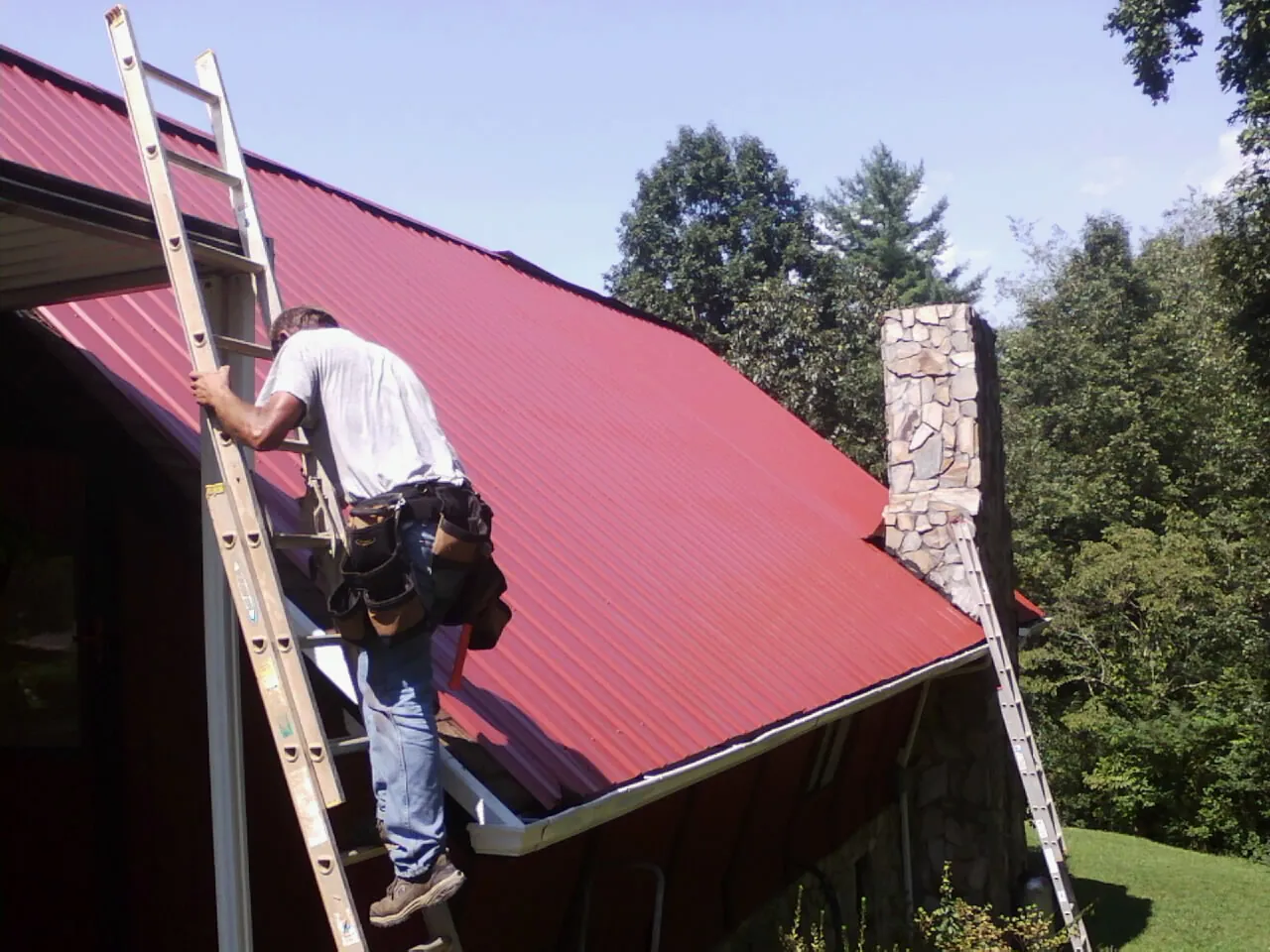 Roofer on a ladder beside a red metal roof and stone chimney — Soffit & Fascia Repair by Roofing Brothers in Hartselle
